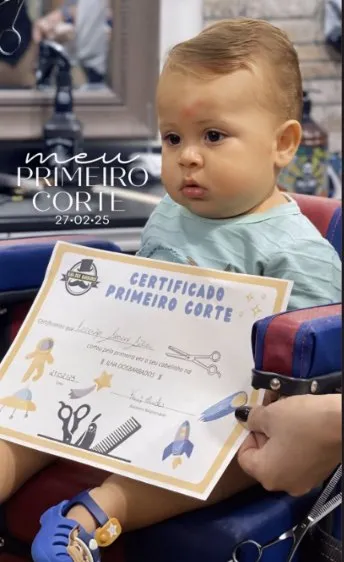 Bebê na cadeira da barbearia segurando o certificado de primeiro corte