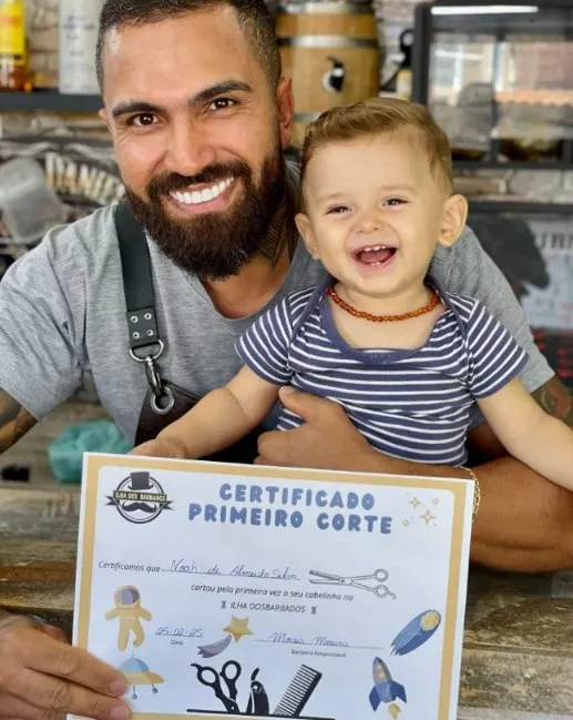 Barbeiro sorrindo com um bebê no colo segurando o Certificado de Primeiro Corte